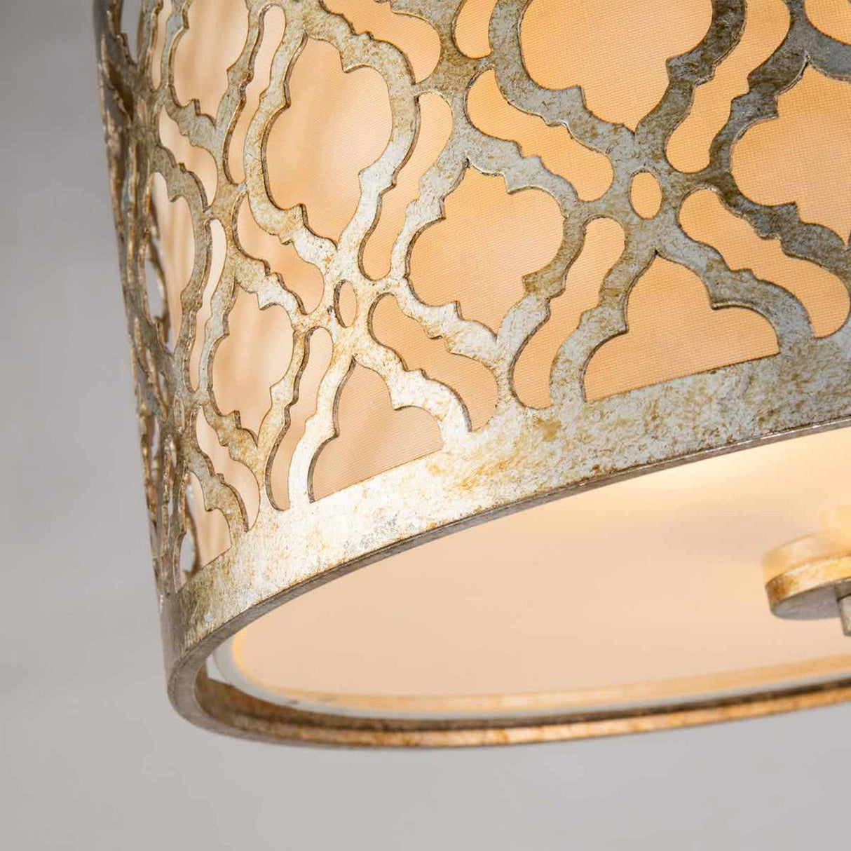 Close-up of the Arabella 2 Light Flush - Gold With Ivory Shade, highlighting a decorative lamp shade with a detailed geometric lattice design in a weathered metallic finish. The distressed gold metal frame permits warm, energy-efficient lighting to pass through its patterned surface.