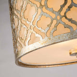 Close-up of the Arabella 2 Light Flush - Gold With Ivory Shade, highlighting a decorative lamp shade with a detailed geometric lattice design in a weathered metallic finish. The distressed gold metal frame permits warm, energy-efficient lighting to pass through its patterned surface.