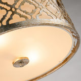 Close-up of the Arabella 2 Light Flush - Gold With Ivory Shade ceiling fixture, showcasing its ornate, distressed gold metal frame adorned with a pattern of curved shapes. The energy-efficient lighting produces a warm glow, accentuating the rustic, weathered finish of the metal.