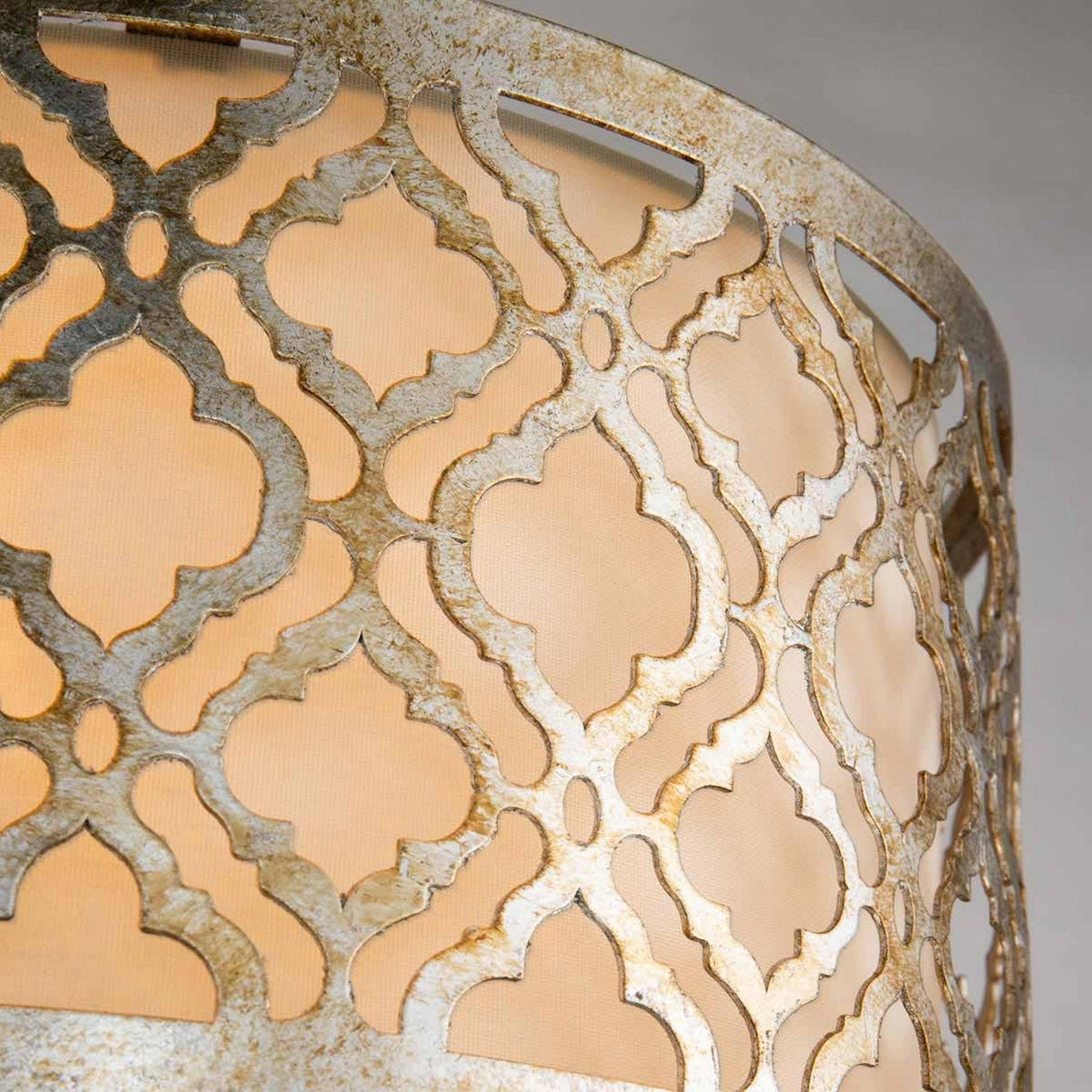 Close-up of the Arabella 2 Light Flush, a lamp that emanates a warm glow with its intricate distressed gold metal lattice design crafted in a Moroccan style, overlaying an ivory fabric shade. The pattern offers a visually appealing contrast against the soft light, showcasing an energy-efficient lighting solution.