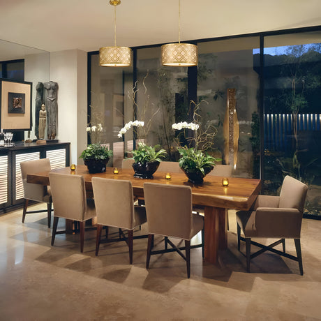 A modern dining room features a wooden table with eight beige chairs, adorned with white orchids and small candles. Two Arabella 3 Light Duo-Mount Large Pendants in gold with ivory shades hang above, while large windows overlook the garden. Nearby, artwork and a sculpture complete the elegant ambiance.