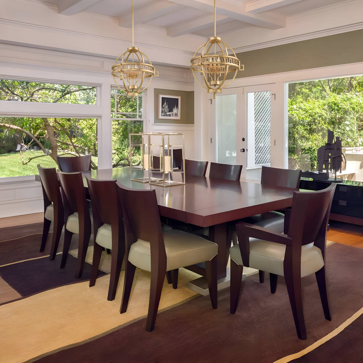 A modern dining room showcases a wooden table encircled by eight upholstered chairs. Large windows provide a view of a lush garden. Above the table, two Lemuria 3 Light Globe Chandeliers with gold finish and ivory shades hang elegantly, featuring adjustable rods. The room is adorned in neutral tones.