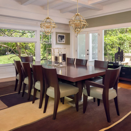 A modern dining room showcases a wooden table encircled by eight upholstered chairs. Large windows provide a view of a lush garden. Above the table, two Lemuria 3 Light Globe Chandeliers with gold finish and ivory shades hang elegantly, featuring adjustable rods. The room is adorned in neutral tones.