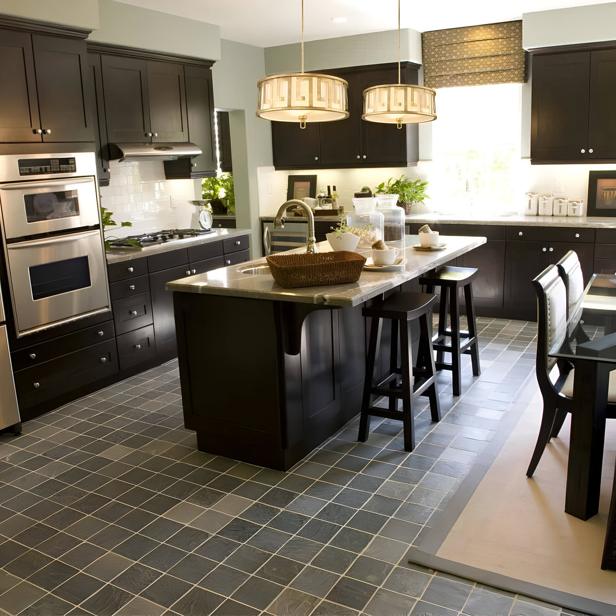 A modern kitchen showcases dark wooden cabinets and an island with bar stools, complemented by stainless steel appliances. Bright natural light streams through a large window, illuminating the Lemuria 3 Light Duo-Mount Large Pendant/Semi-Flush in Gold with an Ivory Shade, featuring adjustable rods above the gray-tiled floor.