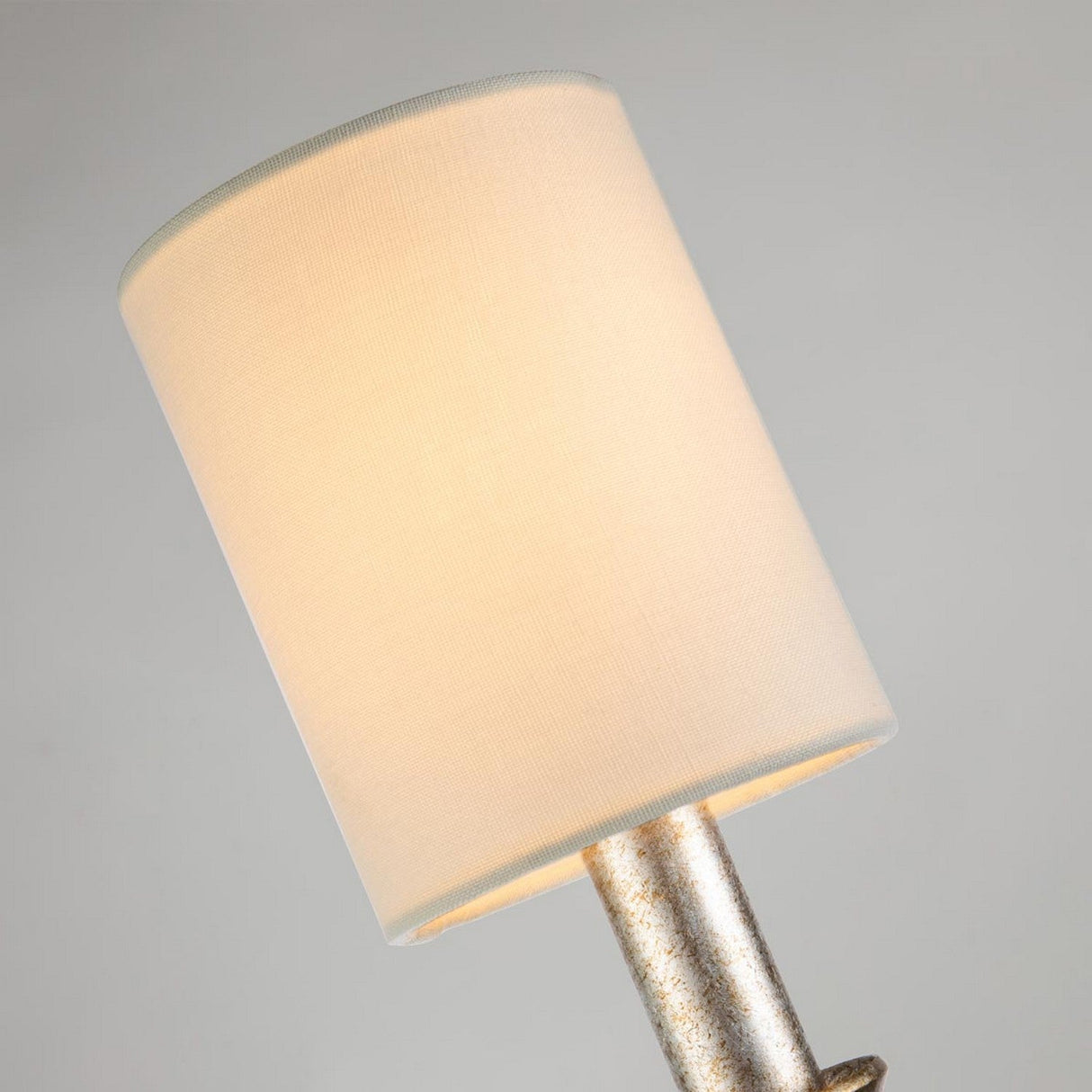 This close-up showcases the Lemuria 1 Light Wall Light, featuring an ivory fabric cylindrical shade. Its metallic base is elegantly highlighted with a gold finish and has a textured design. LED bulbs gently light up the shade, casting a warm glow against a neutral backdrop.