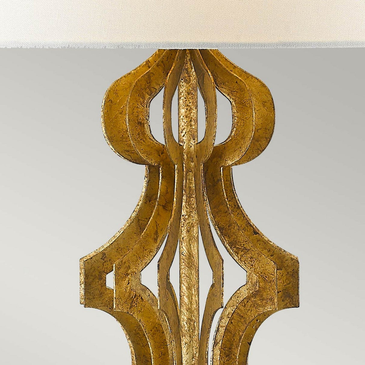 Detailed view of a luxurious gold metallic lamp base showcasing an intricate, symmetrical design, echoing the elegance of the Octavia 1 Light Table Lamp - Gold With Ivory Shade. Set against a light backdrop, the top part of the image displays a portion of an ivory linen shade.