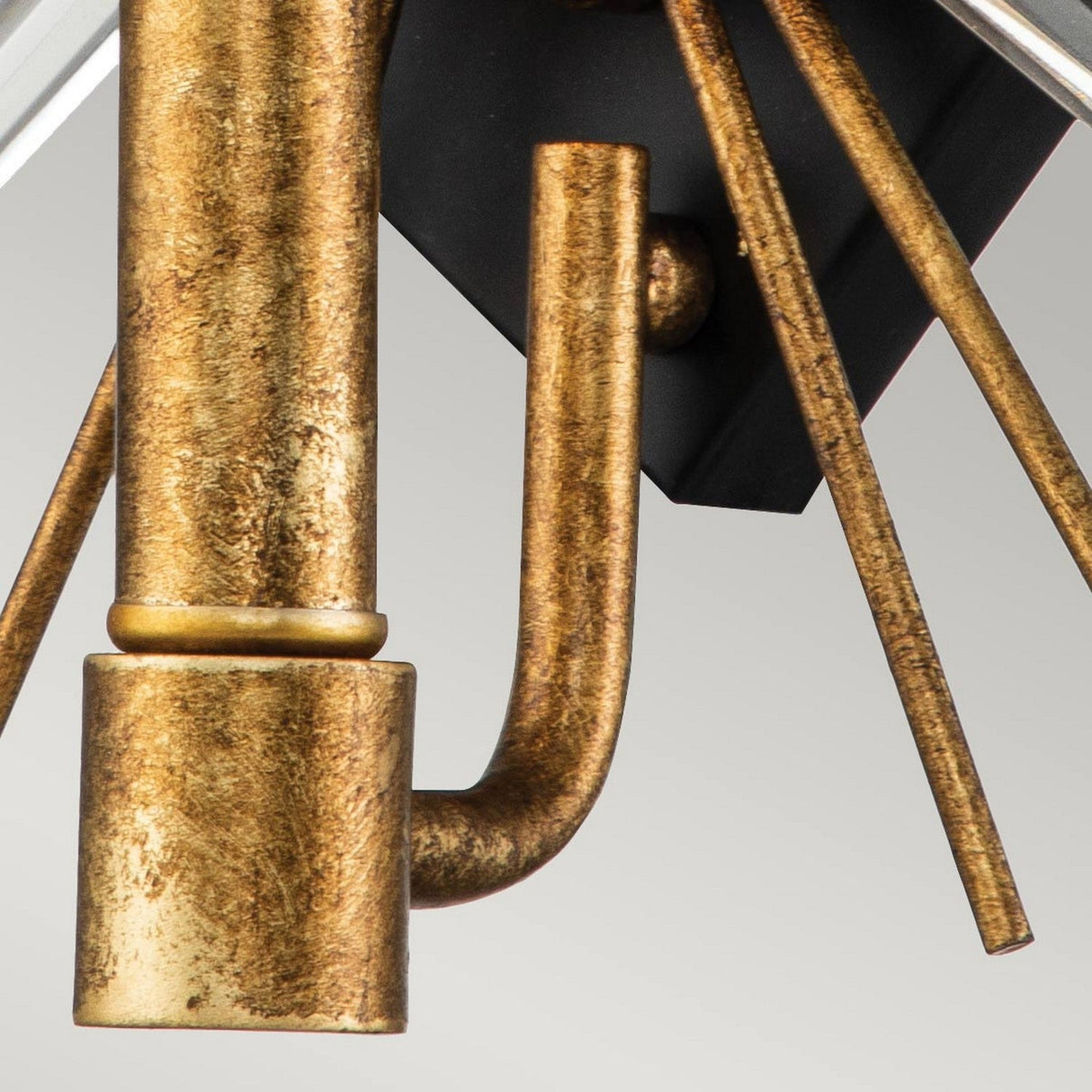 A close-up of the Sun King 1 Light Wall in Gold Leaf, featuring bronze and gold-toned metal with cylindrical shapes and straight rods, artistically arranged against a gray background. The design combines industrial and decorative elements, evoking a modern wall light.