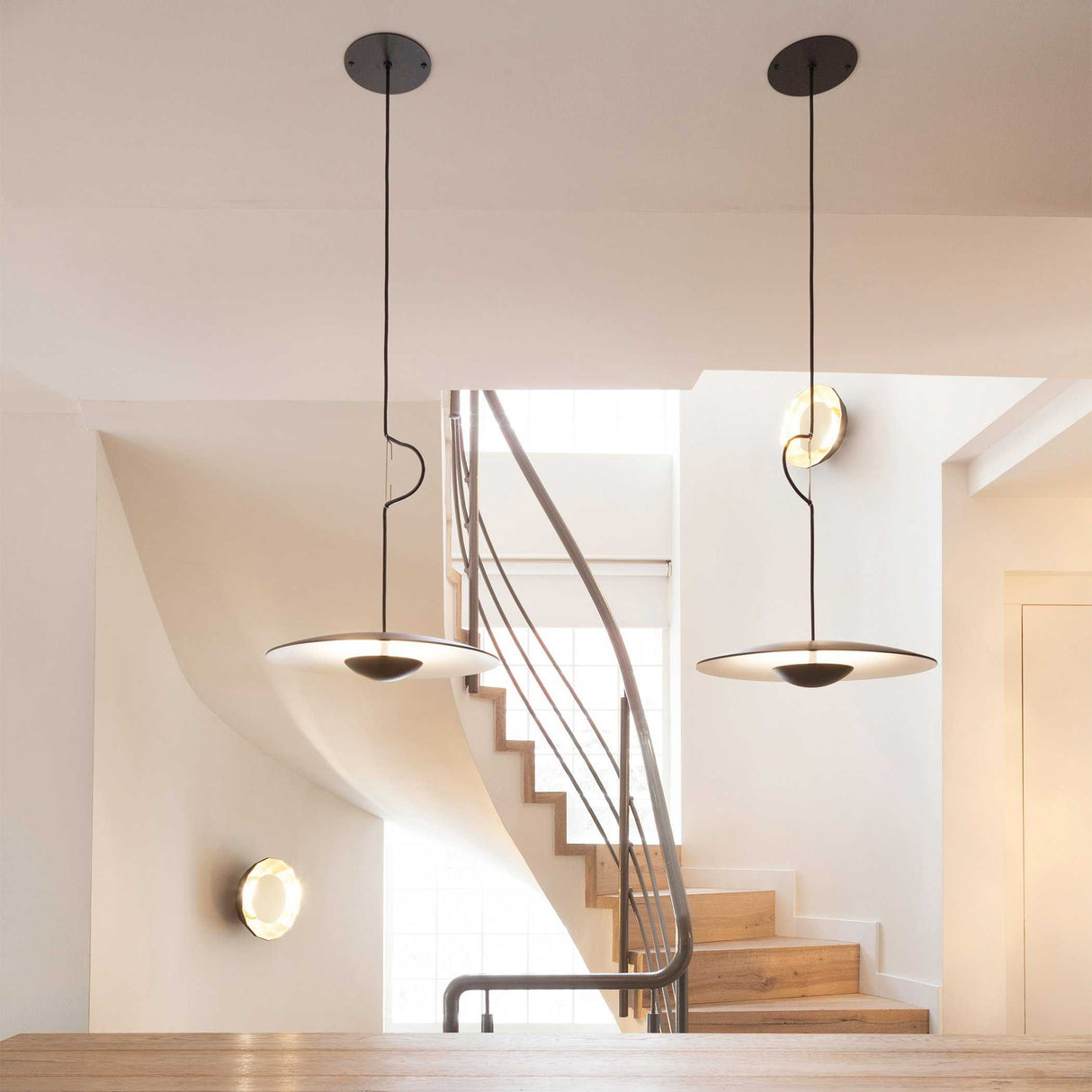 The Ginger LED Pendant Light in Black & Oak complements a minimalist design alongside a floating staircase with wooden steps. Soft, natural light filters through a window, creating an airy atmosphere. The white walls accentuate the clean, eco-conscious aesthetic, highlighting the modern charm.