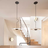 The Ginger LED Pendant Light in Black & Oak complements a minimalist design alongside a floating staircase with wooden steps. Soft, natural light filters through a window, creating an airy atmosphere. The white walls accentuate the clean, eco-conscious aesthetic, highlighting the modern charm.