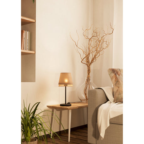 Golanne Table Lamp in black steel with a tapered beige fabric shade and wooden rod fringe, shown lit on a wooden side table beside a sofa.