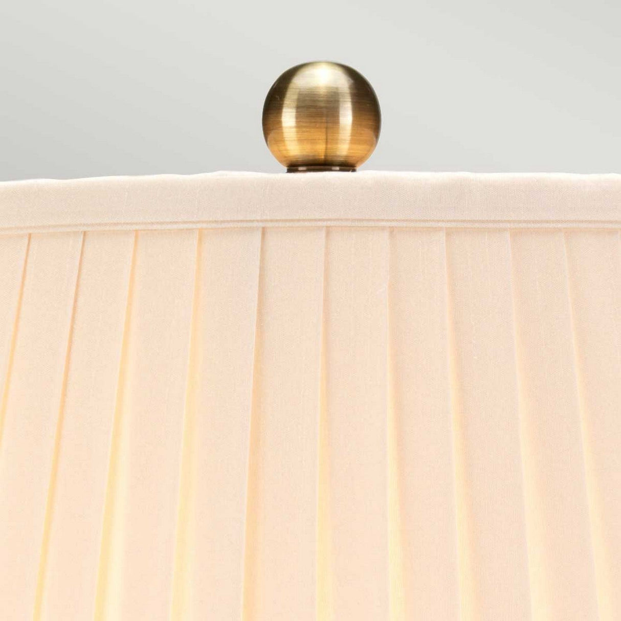 Close-up of a beige pleated lampshade with a brass finial on top. The spherical finial gleams, contrasting the soft fabric shade. Below, the Gold Birds 1 Light Table Lamp in light blue and gold sits elegantly, its Chinese porcelain base lending a touch of classic artistry to the off-white backdrop.