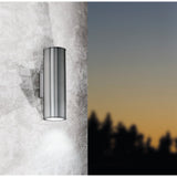 Gorbora LED 2-light outdoor wall light in stainless steel, slim cylindrical design mounted on a textured wall, shown at dusk casting up and down light.