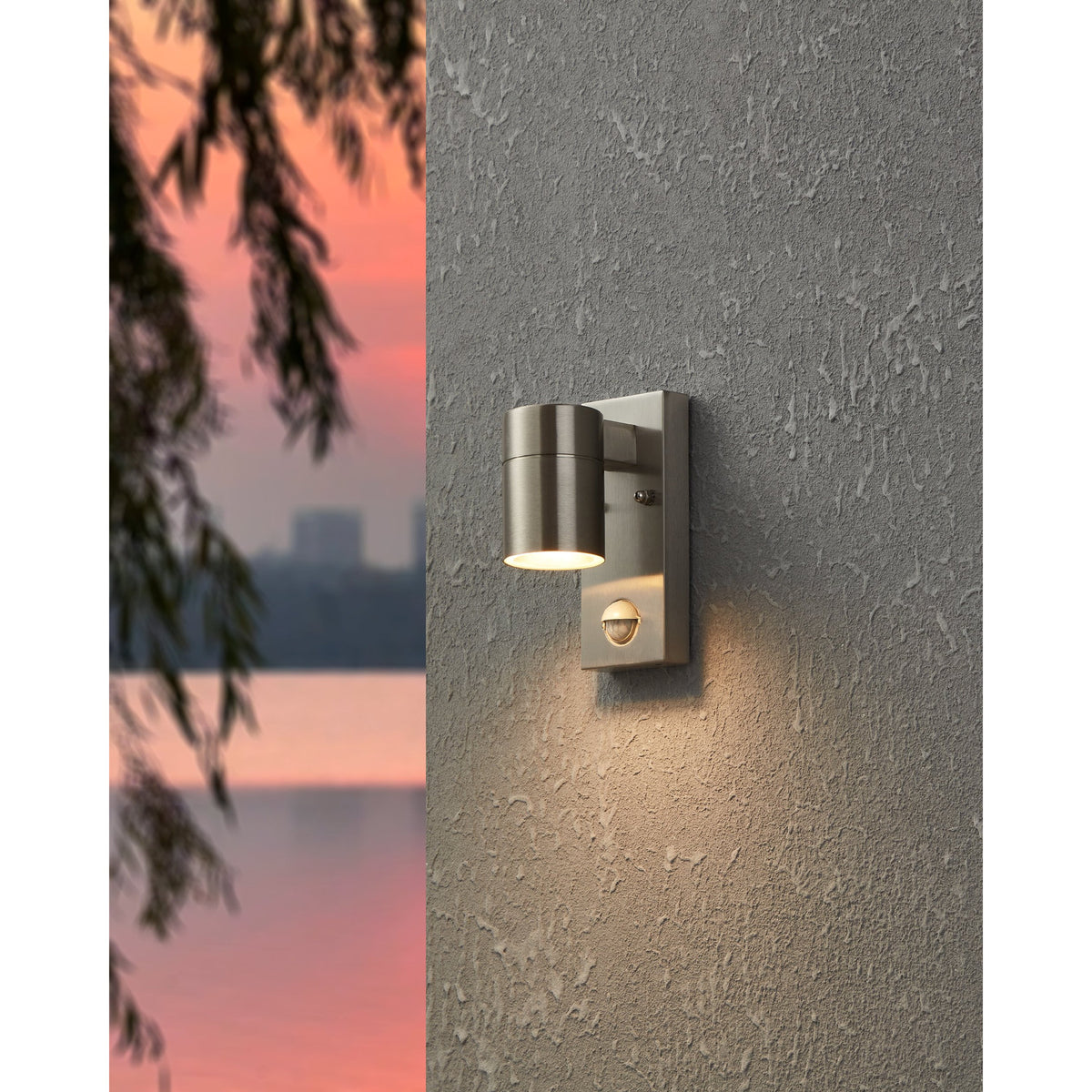 Gorbora outdoor wall light in stainless steel, cylindrical head with motion sensor, mounted on a grey textured wall and illuminated at dusk.