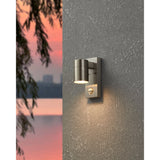 Gorbora outdoor wall light in stainless steel, cylindrical head with motion sensor, mounted on a grey textured wall and illuminated at dusk.