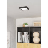 Gosmelle LED Downlight 15 x 15cm IP20 in Black, a square surface-mounted ceiling fixture with black plastic frame and white diffuser, shown above a bookshelf in a living room setting.