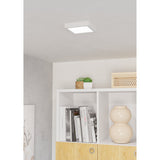 Gosmelle square LED downlight in white plastic, 15 x 15cm, mounted flush on a ceiling above open shelving with books and decor in a living room setting.