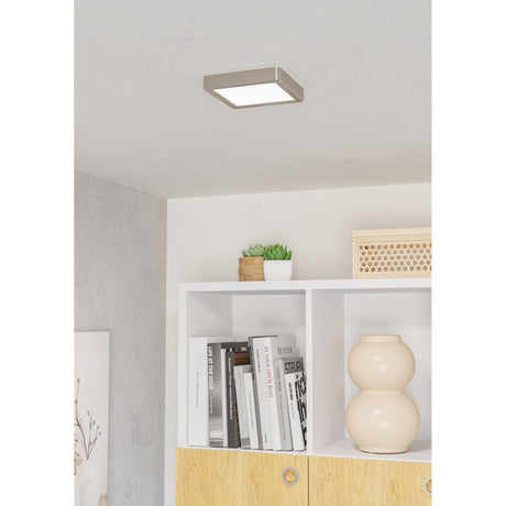 Gosmelle LED Downlight 15 x 15cm in satin nickel with a square frame and white diffuser, shown fitted to a ceiling above a white bookshelf with books, a vase and plants.