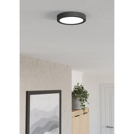 Gosmelle round LED downlight in black plastic with white diffuser, surface-mounted on a ceiling in a hallway setting with artwork and plant below.