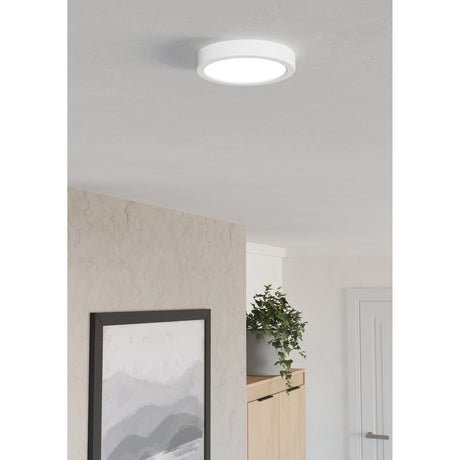 Gosmelle round LED downlight in white plastic, 16.6 x 2.5cm, surface-mounted on a ceiling in a neutral room with a plant and framed artwork below.