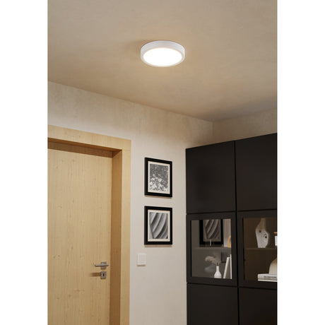 Gosmelle LED Downlight, 21.1cm round white plastic ceiling fixture with flat diffuser, shown mounted on a hallway ceiling near a wooden door, framed artwork, and black cabinetry.