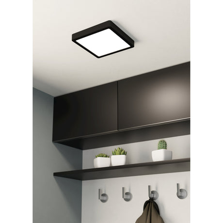 Gosmelle LED downlight with black steel frame and white plastic diffuser mounted on a hallway ceiling above dark cabinets and a shelf with potted plants.