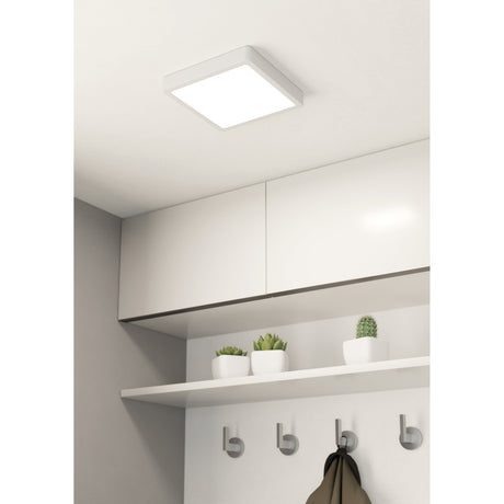 Gosmelle square LED downlight, 21 x 21cm white steel body with flat white diffuser, fitted on a hallway ceiling above white wall cabinets and a shelf with potted plants.