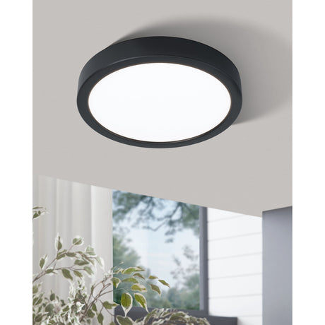 Gosmelle LED Downlight, 21cm round black steel ceiling fixture with white diffuser, shown glowing warm white in a room ceiling corner near a window and plant.