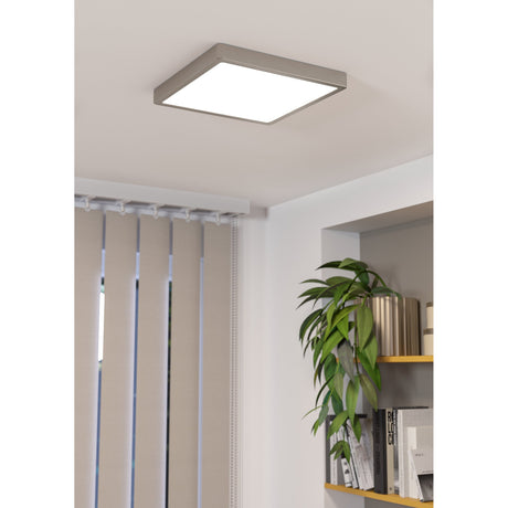Square LED downlight with slim satin nickel frame and flat white diffuser, mounted on a living room ceiling near shelving and vertical blinds.