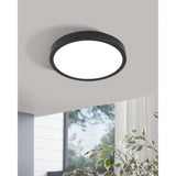 Gosmelle LED Downlight 28.5cm in black steel with a round white plastic diffuser, mounted flush on a grey ceiling in a room with a window and plant visible below.