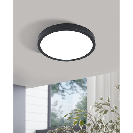Gosmelle LED Downlight 28.5cm in black steel with a round white plastic diffuser, mounted flush on a grey ceiling in a room with a window and plant visible below.