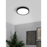 Gosmelle 28.5cm round LED downlight in black with white diffuser, shown flush-mounted on a white ceiling in a grey living room with artwork and a plant below.