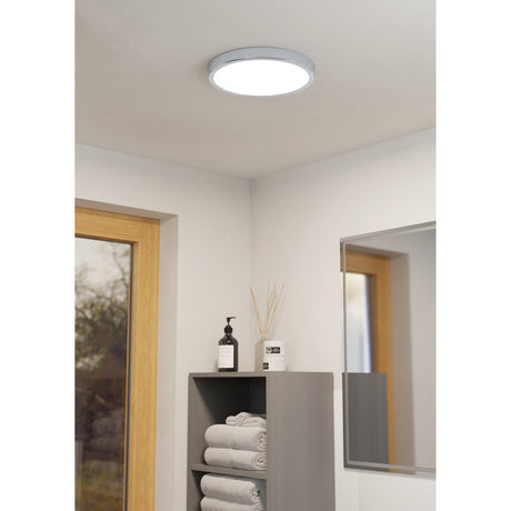 Gosmelle round LED downlight in chrome with white diffuser, flush-mounted on a bathroom ceiling above a grey storage unit with towels and toiletries.
