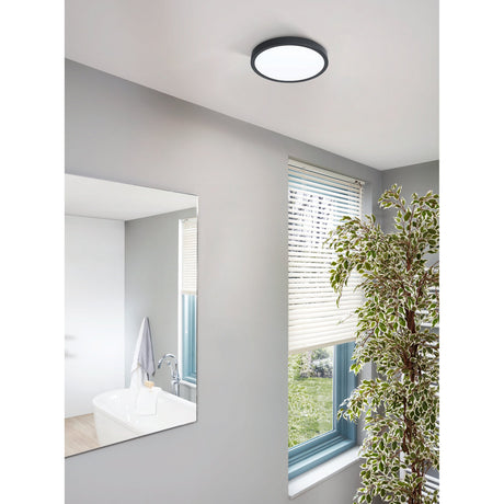 Gosmelle round LED downlight with black rim and white diffuser fitted to a bathroom ceiling, shown above a window and mirror reflecting a sink.