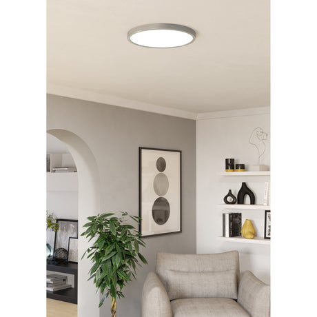 Round LED downlight in satin nickel with white diffuser, fitted flush to a light grey ceiling above a neutral living room with armchair, framed art and shelving.