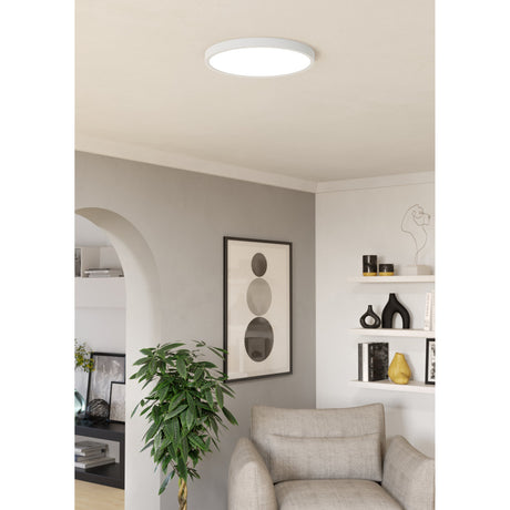 Gosmelle LED Downlight 38.9cm in white, a slim round flush ceiling fixture shown illuminating a living room with an armchair, framed art, and open shelving.