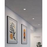 Gosmelle round LED downlight in white recessed flush into a hallway ceiling, shown alongside framed artwork and a plain light grey wall.