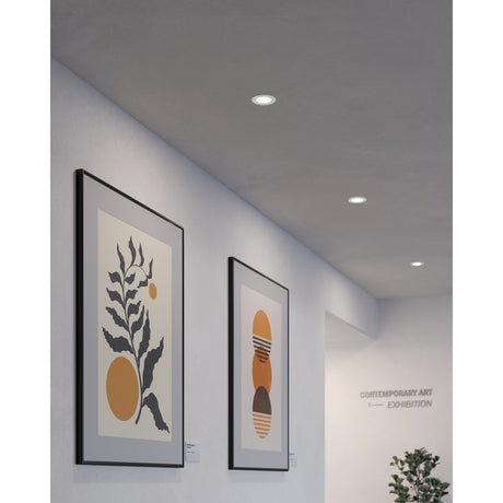 Gosmelle round LED downlight in white recessed flush into a hallway ceiling, shown alongside framed artwork and a plain light grey wall.
