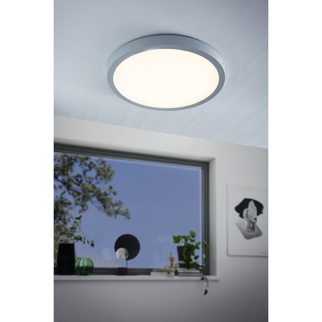 Gosmelle LED Downlight in silver, a round ceiling fixture with a white diffuser, shown illuminated on a white ceiling in a room with a window and plants.