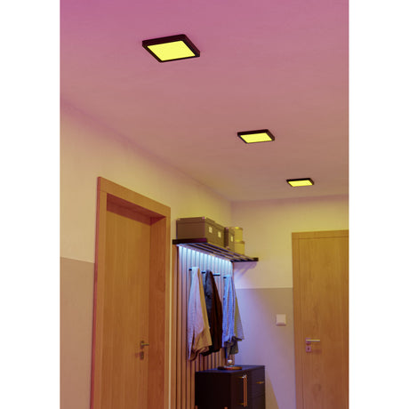 Three square black-framed Gosmelle LED downlights recessed into a pink-toned hallway ceiling, each glowing warm yellow above a coat rack and wooden doors.