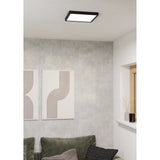 Gosmelle square LED downlight, 29 x 29cm, with black plastic frame and white diffuser, mounted flush on a living room ceiling above a grey wall with framed art and a green sofa.