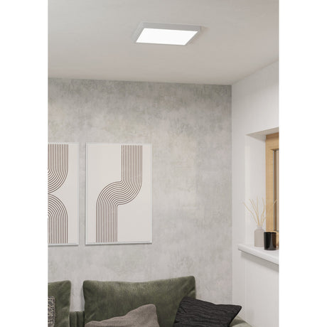 Gosmelle square LED downlight in white plastic, flush-mounted on a living room ceiling above a grey textured wall with framed line-art prints and a green sofa.