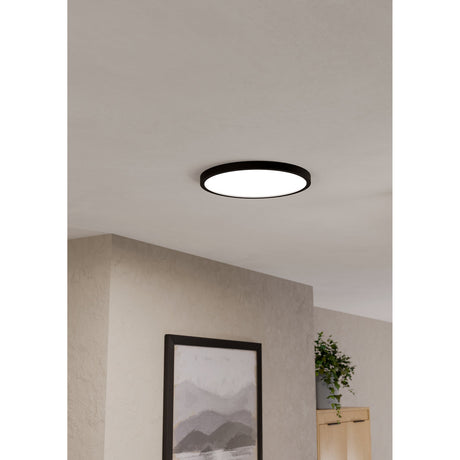 Gosmelle Smart LED Downlight 38.9cm in black, a slim round surface-mounted ceiling light with white diffuser, shown illuminated in a neutral hallway near artwork and a plant.