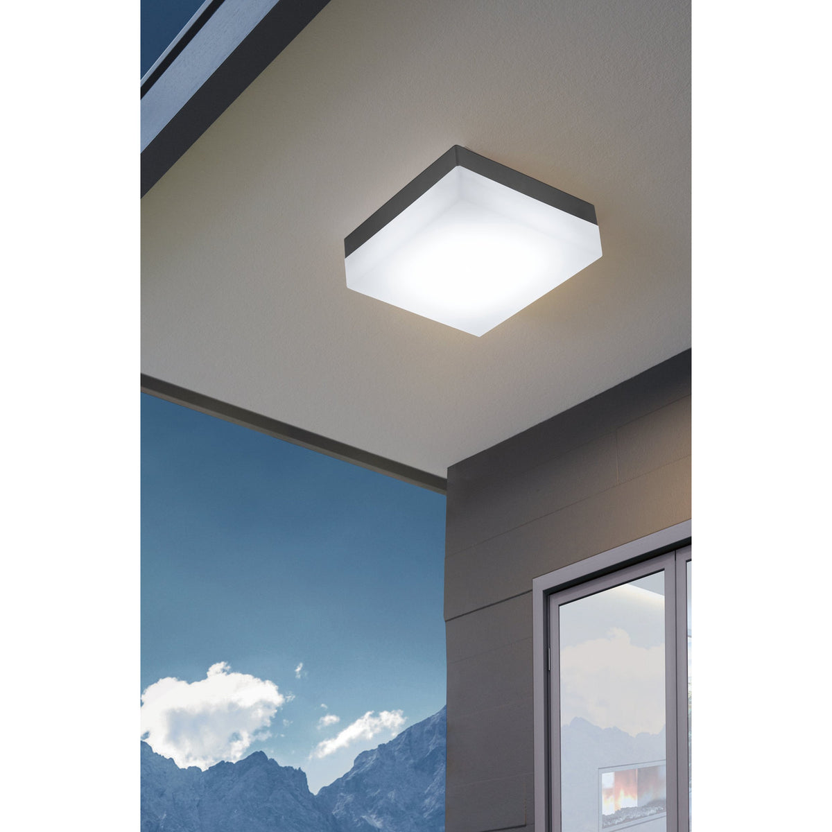Gramane LED outdoor wall light with square anthracite plastic body and white diffuser, shown mounted on a ceiling soffit outside a modern home with mountain view.
