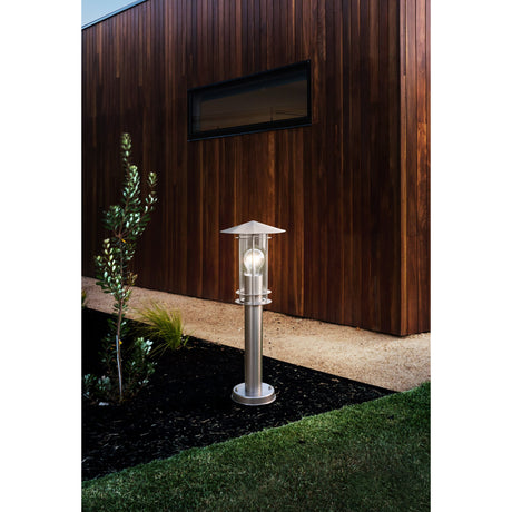 Granette Bollard Light in stainless steel with a conical lantern hat over a clear glass shade, illuminated on a post beside a garden path and timber-clad house exterior.