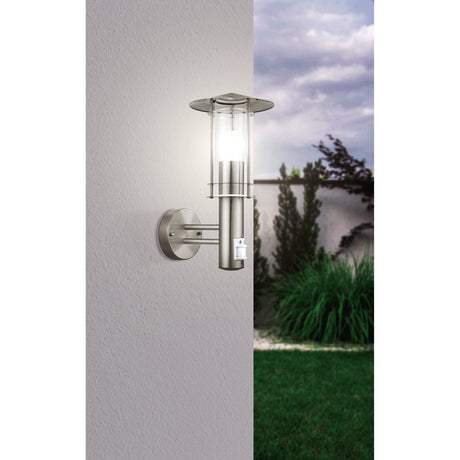 Granette outdoor wall light in stainless steel with clear glass cylinder, hat-shaped cap and motion sensor, mounted on a grey exterior wall beside a garden view.
