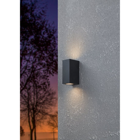 Granvenne 2 Light Outdoor Wall Light in black, rectangular design mounted on a textured grey wall, casting upward and downward beams, shown at dusk beside silhouetted trees.