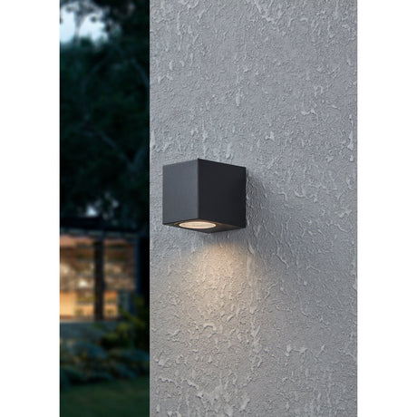 Granvenne Outdoor Wall Light in black, a compact box-shaped plastic fixture mounted on a textured grey exterior wall, casting downward light from its round lens.