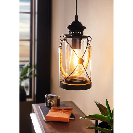 Grasmenne clear glass pendant light with black steel lantern frame, crossed bars, and rope handle, shown hanging above a desk with books and a plant.