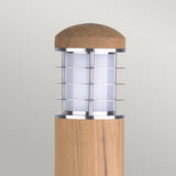 The Poole 1 Light Bollard in light brown showcases a contemporary outdoor design with a cylindrical Malaysian teak base and a Marine Grade 316 stainless steel metal cage surrounding a white frosted glass shade. It is finished with a wooden dome and is set against a simple, light gray background.