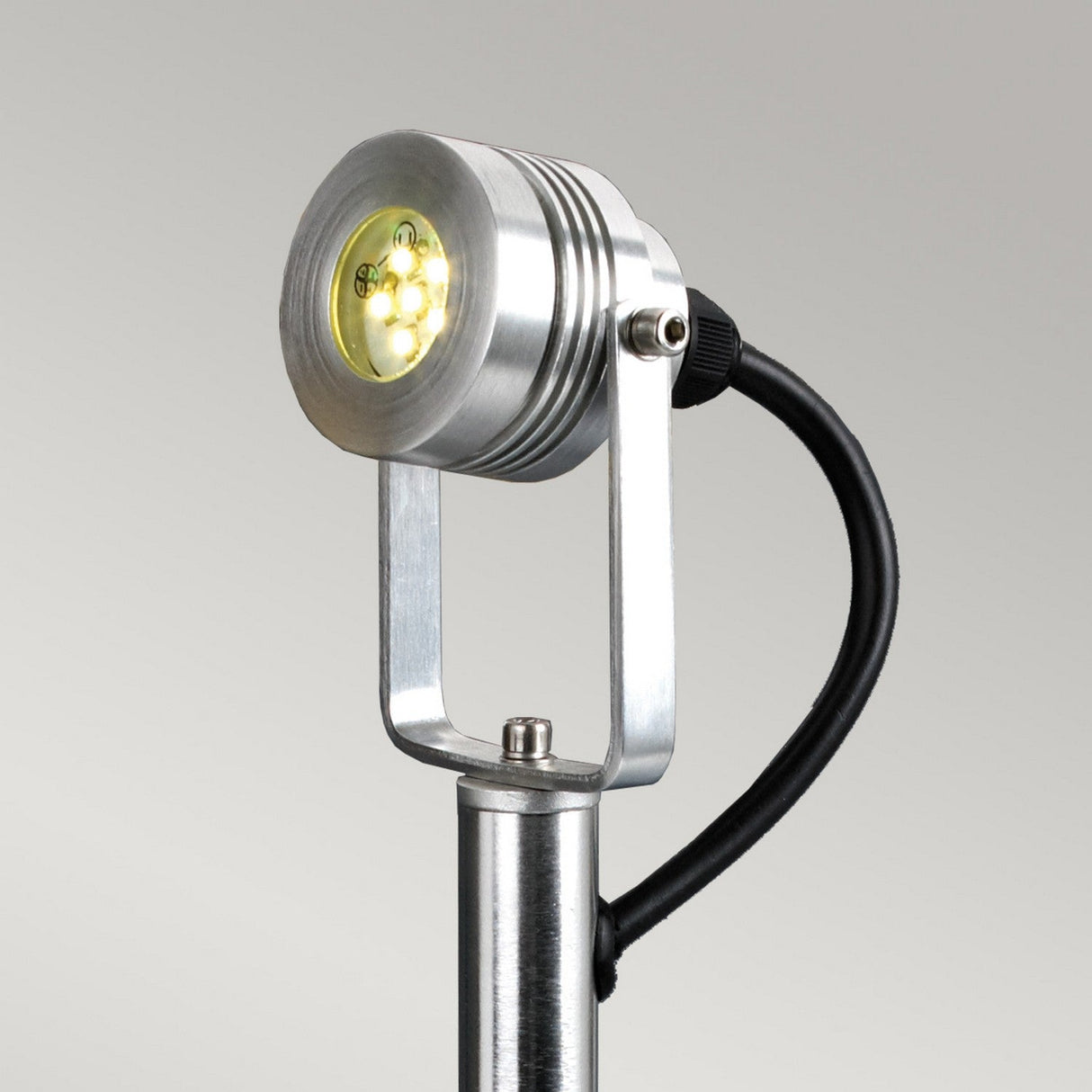 A detailed view of the Spennymoor 1 x 12V Spotlight and Pole in a sleek silver finish, featuring a circular head with an array of small LED bulbs. The low voltage fixture is mounted on a pole with a 1-meter black cable extending from the rear, set against a simple light gray background.